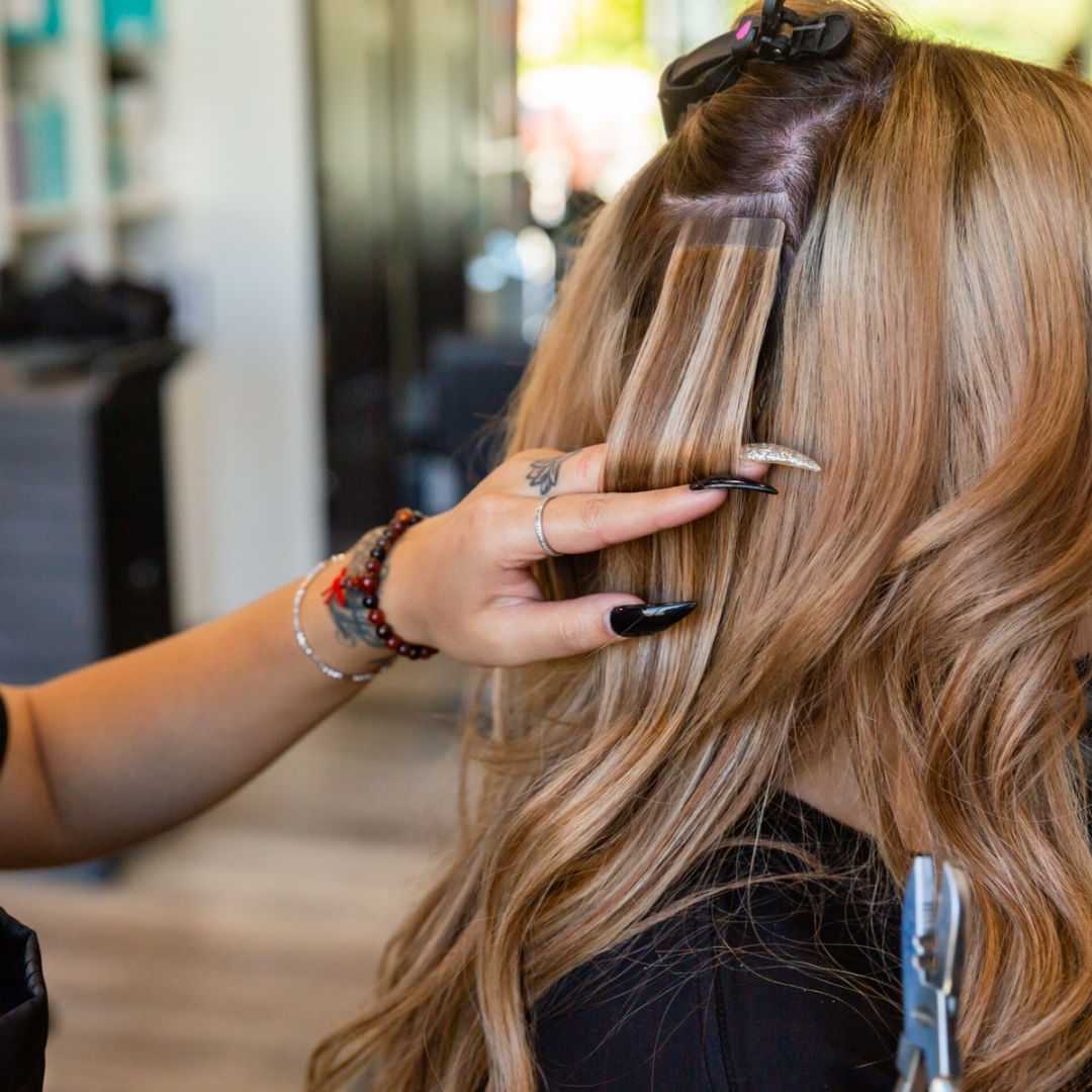 Hair salon stylist applies extensions on woman's blonde hair.